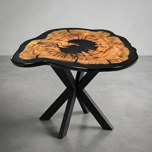 May include: A table featuring a natural wood slab tabletop with a live edge, encased in a smooth black resin border. The center of the wood displays visible grain patterns and a dark void. The base consists of four black metal legs in an X-shape.
