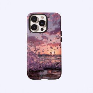 May include: A rectangular smartphone case featuring a printed image of a sunset over the ocean with a prominent water splash in the foreground. The design displays shades of purple, pink, and orange, with clear water droplets scattered across the surface.
