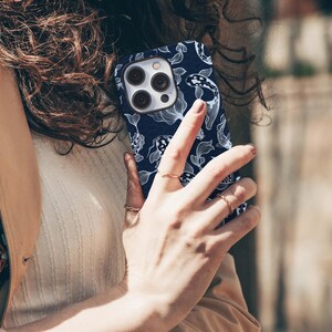 May include: A navy blue phone case featuring a white koi fish and aquatic plant pattern. The case has a camera cutout and is held in a person's hand. The design is a repeating pattern of koi fish and aquatic plants.