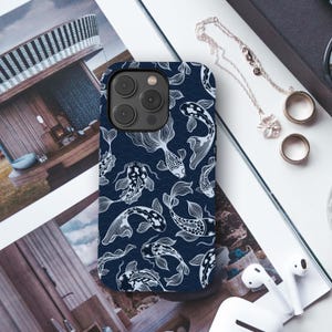 May include: A navy blue phone case featuring a white koi fish pattern. The design includes stylized koi fish and wave patterns. The case is shown on a magazine with jewelry and white earbuds.