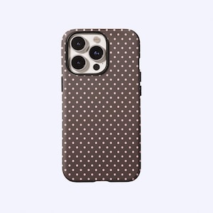 Puede incluir: Una funda de teléfono marrón con un patrón de pequeños lunares rosas espaciados uniformemente. La funda tiene un borde oscuro alrededor de las lentes de la cámara y un marco negro. La funda está diseñada para un smartphone.