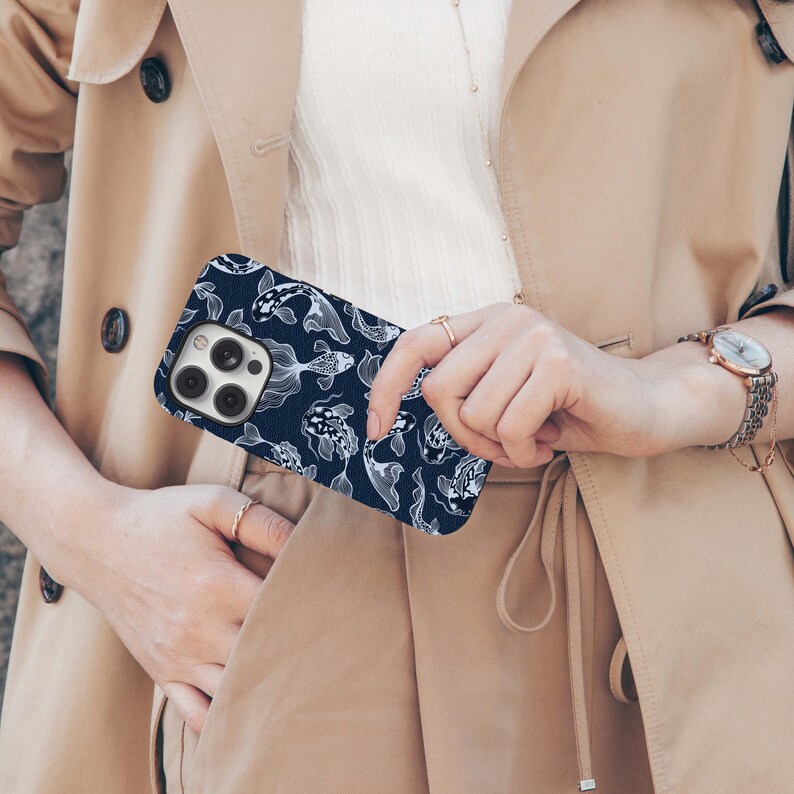 May include: A navy blue phone case featuring a white koi fish pattern. The case is held in front of a beige trench coat. The phone case has a camera cutout and button placements.