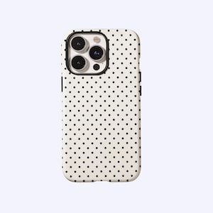 Peut inclure: Une coque de téléphone blanche avec un motif à pois noirs. L'étui a un bord noir autour de l'appareil photo et un cadre noir. La coque est conçue pour s'adapter à un smartphone.
