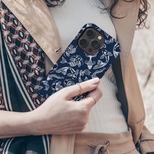 May include: A navy blue phone case featuring a white koi fish pattern. The case has a matte finish and a camera cutout. The phone case is held by a person wearing a beige jacket and a white top.