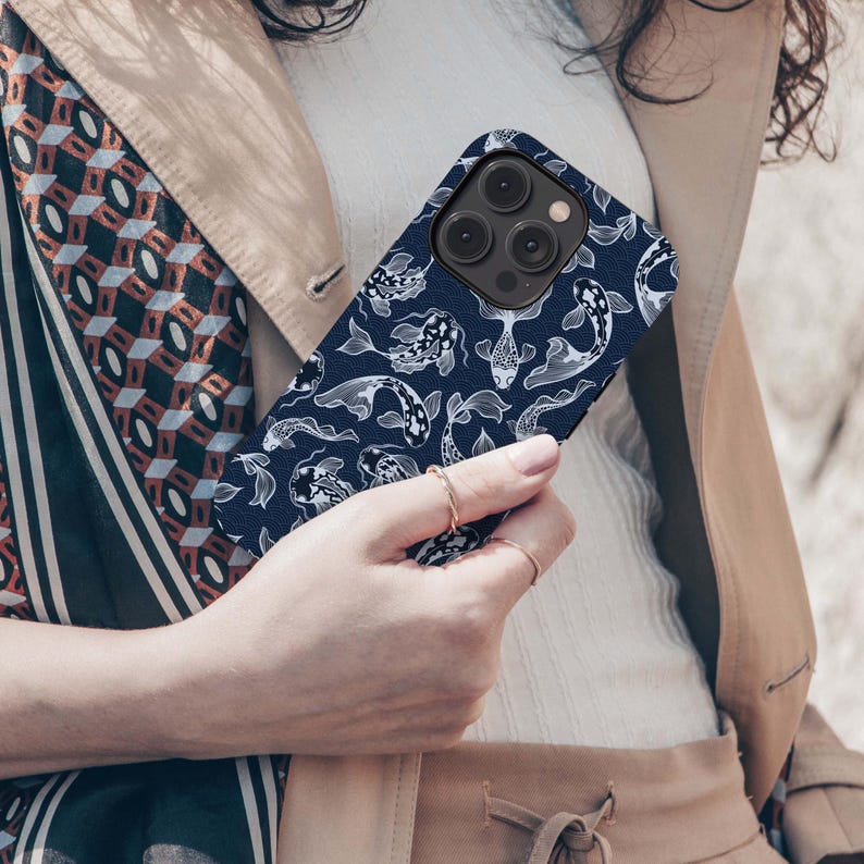 May include: A navy blue phone case featuring a white koi fish pattern. The case has a matte finish and a camera cutout. The phone case is held by a person wearing a beige jacket and a white top.