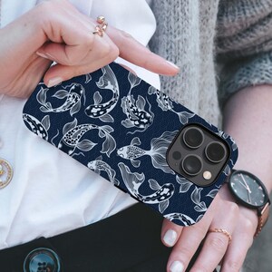 May include: A navy blue phone case featuring a white koi fish pattern. The design includes detailed illustrations of koi fish and wave patterns. The case has a black camera module and is held by a person.