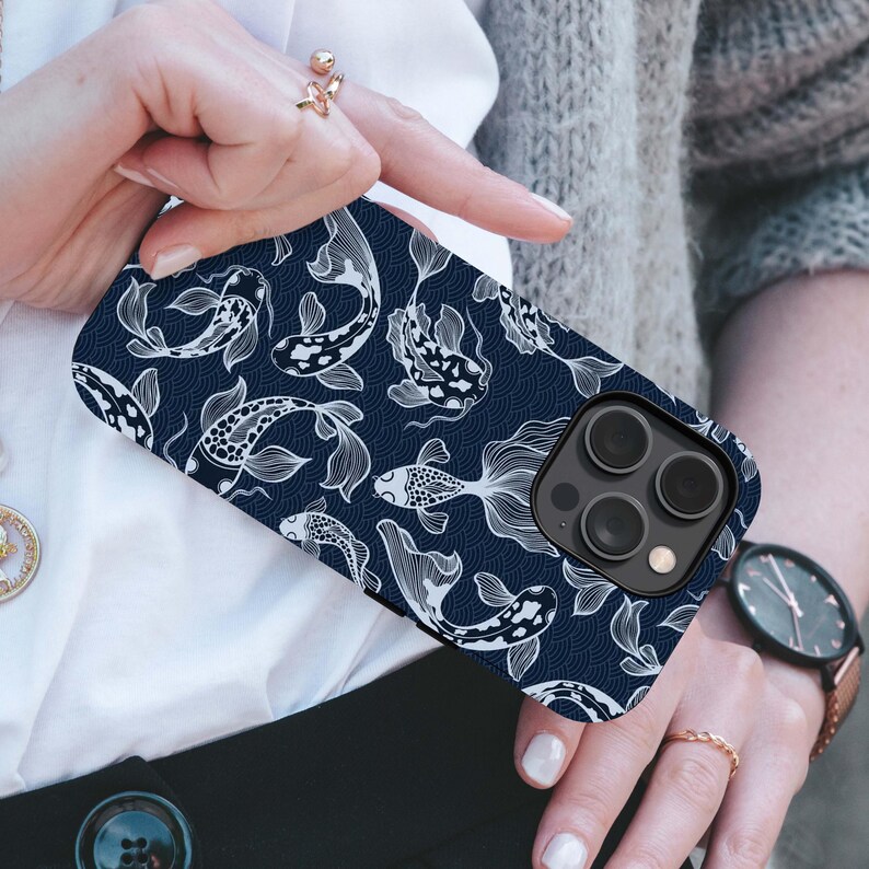May include: A navy blue phone case featuring a white koi fish pattern. The design includes detailed illustrations of koi fish and wave patterns. The case has a black camera module and is held by a person.