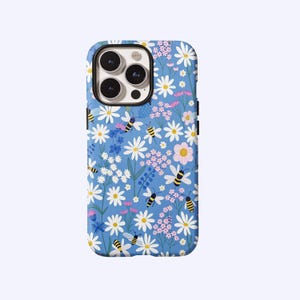 May include: A blue phone case with a floral design of white daisies, pink flowers, and yellow and black bees. The case has a glossy finish and a camera cutout.