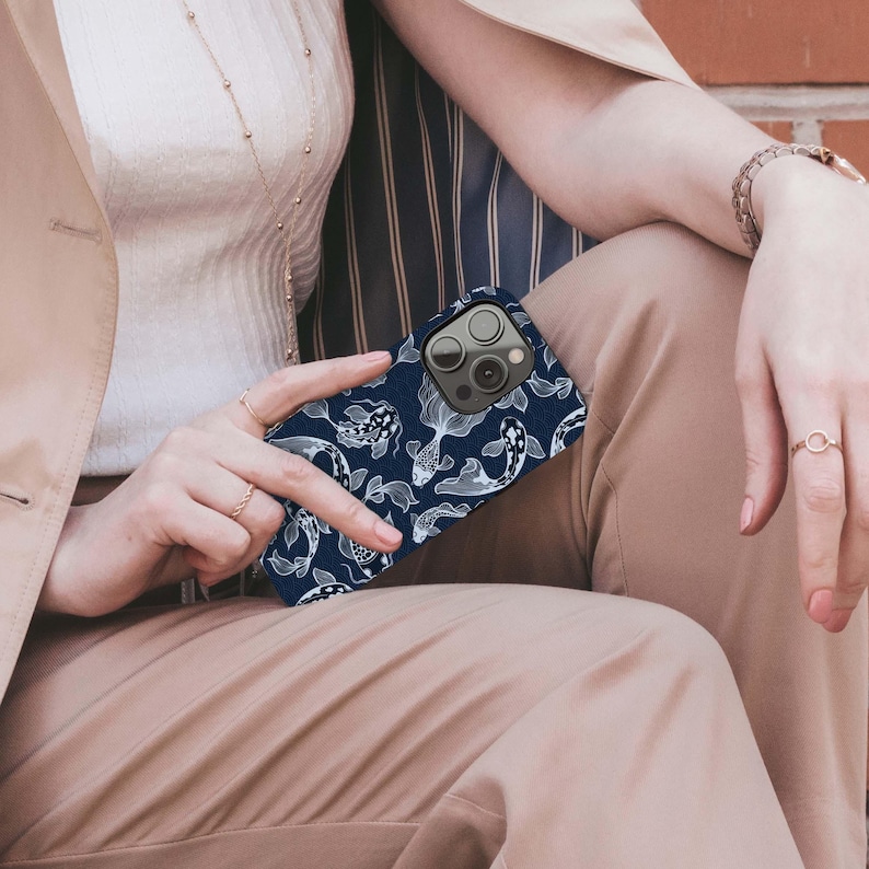 May include: A navy blue phone case featuring a white koi fish pattern. The case has a protective design and a camera cutout. The phone case is held by a person wearing rings and a bracelet.