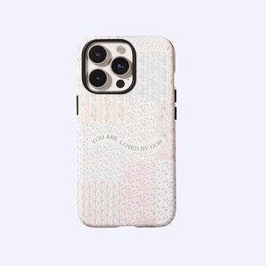 Puede incluir: Funda de teléfono con diseño de patchwork floral y el texto "YOU ARE LOVED BY GOD". La funda presenta una mezcla de patrones florales en rosa pastel, azul y blanco. Tiene un borde negro para la lente de la cámara y botones laterales negros.