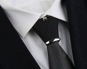 May include: A close-up shot of a black tie with a textured pattern, held in place by a silver tie clip and a silver tie bar. The tie is worn with a white shirt and a black suit jacket.