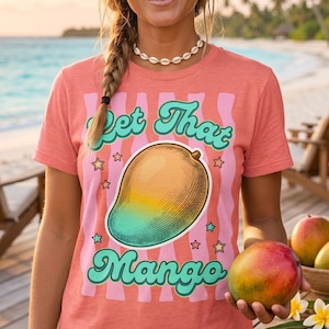 以下が含まれることがあります： 「Let That Mango」の文字とマンゴーのイラストがプリントされたコーラル色のTシャツ。熟したマンゴーを持っています。背景にはヤシの木と夕焼けのビーチシーンが描かれています。