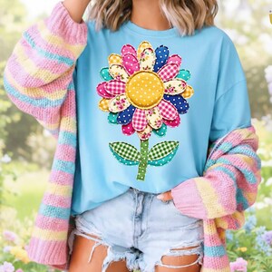 Könnte beinhalten: Hellblaues T-Shirt mit einem großen, bunten Blumenmuster. Die Blume hat eine gelbe Mitte und Blütenblätter in verschiedenen Mustern und Farben. Die Person trägt eine gestreifte Strickjacke in Pastellfarben und Jeansshorts.