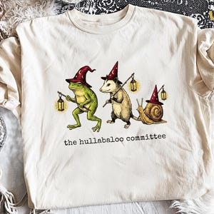 May include: A cream-coloured t-shirt featuring a graphic of a green frog, an opossum, and a snail, each wearing a maroon wizard hat with moon and star symbols. The frog and opossum carry lanterns on sticks. The text reads, the hullabaloo committee.