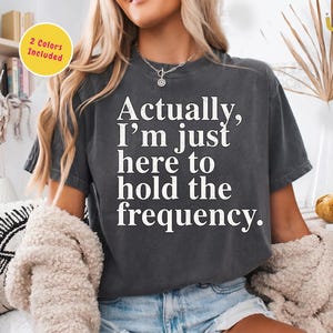 Op de afbeelding: Donkergrijs t-shirt met de witte tekst: "Eigenlijk ben ik er alleen maar om de frequentie vast te houden." Het ontwerp is voor sublimatie/DTF-druk, met 2 kleuren en een transparante achtergrond.