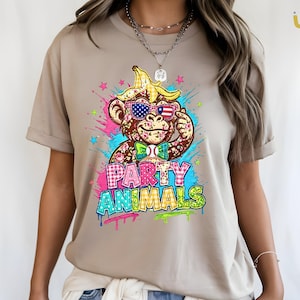 Puede incluir: Camiseta color beige con un gr&aacute;fico colorido de un mono con gafas de sol, un sombrero de c&aacute;scara de pl&aacute;tano y una pajarita. El texto "PARTY ANIMALS" se muestra en una fuente de estilo graffiti con efectos de salpicaduras de pintura.