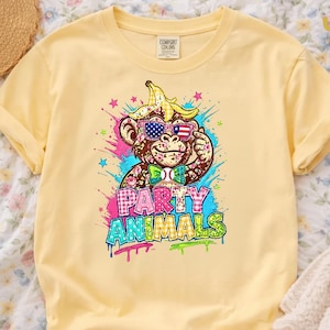 Puede incluir: Camiseta amarilla clara con un mono de dibujos animados que lleva gafas de sol con un dise&ntilde;o de bandera estadounidense y un pl&aacute;tano en la cabeza. La camiseta tiene las palabras "PARTY ANIMALS" en letras coloridas de estilo grafiti.