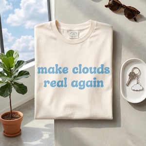 Op de afbeelding: Crèmekleurig T-shirt met de tekst "make clouds real again" in lichtblauwe letters. Het shirt is opgevouwen op een lichtgrijze ondergrond, naast een zonnebril en een klein schaaltje met sleutels.