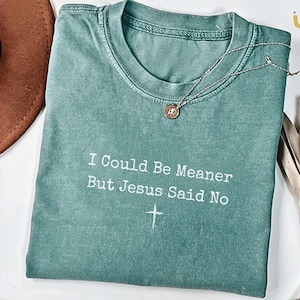 May include: A teal-colored t-shirt with the text "I Could Be Meaner But Jesus Said No" and a cross graphic. A silver necklace with a pendant rests on the shirt. A brown hat is partially visible in the background.
