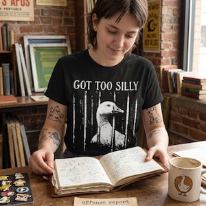 May include: Black t-shirt with the text "GOT TOO SILLY" and a graphic of a goose behind bars. The image includes a mug with a goose design, a notebook, handcuffs, and a dollar bill.