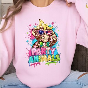 Puede incluir: Sudadera rosa claro con un mono de dibujos animados con gafas de sol con un dise&ntilde;o de bandera estadounidense y una c&aacute;scara de pl&aacute;tano en la cabeza. Las palabras "PARTY ANIMALS" se muestran en letras de estilo graffiti coloridas.