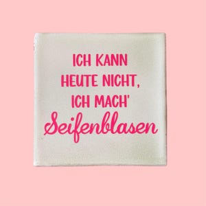 Könnte beinhalten: Eine quadratische, cremefarbene dekorative Fliese mit dem deutschen Satz "ICH KANN HEUTE NICHT, ICH MACH' Seifenblasen" in leuchtend rosa Schrift. Die Fliese ist vor einem zartrosa Hintergrund platziert und erzeugt eine verspielte und skurrile Ästhetik.