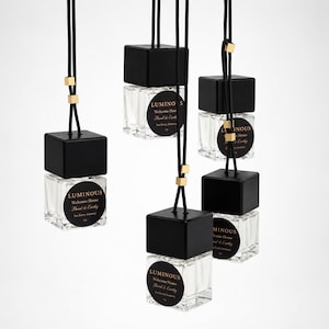 May include: Five square glass car air freshener bottles with black wooden caps, hanging from black cords with small gold-toned metal beads. Each bottle features a round black label with gold text reading Luminous, Welcome Home, Floral & Earthy, For Every Journey.