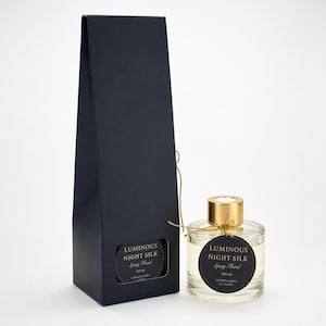 May include: A tall, dark navy blue rectangular gift box stands next to a clear glass perfume bottle with a gold cap. Both feature circular black labels with gold text reading Luminous Night Silk, Spicy Floral, 100 ml, London Born UK Based.
