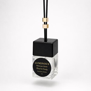 May include: A square glass car air freshener bottle with a black cap and a black hanging cord featuring two gold-toned metal beads. A circular black label on the front reads: LUMINOUS, Welcome Home, Floral & Earthy, For Every Journey, UK.
