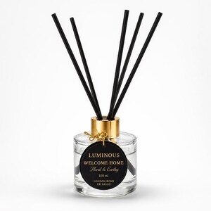 May include: A clear glass reed diffuser bottle with a gold collar and five black reeds. A round black label features gold text reading LUMINOUS WELCOME HOME Floral & Earthy 100 ml LONDON BORN UK BASED. A thin gold string attaches the label to the neck.