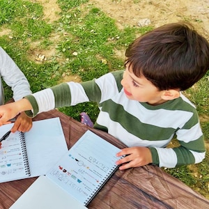 Puede incluir: Un ni&ntilde;o con una camiseta a rayas verdes y blancas mira un cuaderno de espiral. El cuaderno abierto revela texto manuscrito e ilustraciones coloridas. Otra persona tambi&eacute;n est&aacute; trabajando en un cuaderno similar.