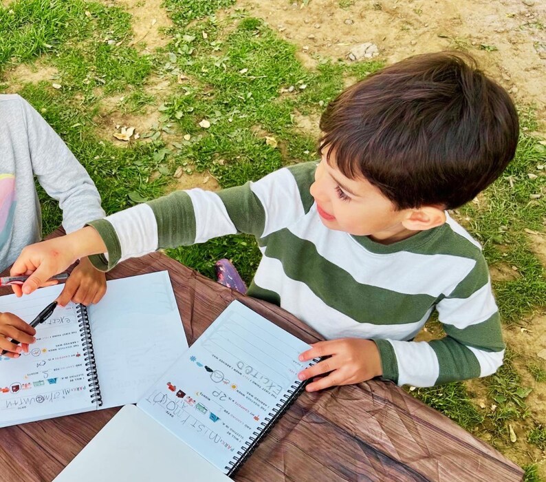 Puede incluir: Un ni&ntilde;o con una camiseta a rayas verdes y blancas mira un cuaderno de espiral. El cuaderno abierto revela texto manuscrito e ilustraciones coloridas. Otra persona tambi&eacute;n est&aacute; trabajando en un cuaderno similar.
