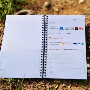 Puede incluir: Diario de camping con espiral abierto, con p&aacute;ginas rayadas y elementos decorativos. La p&aacute;gina izquierda est&aacute; en blanco, mientras que la p&aacute;gina derecha presenta indicaciones para registrar experiencias de camping. El diario est&aacute; abierto sobre una superficie natural.