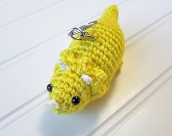 May include: A yellow crocheted dinosaur keychain with black eyes and white horns. The keychain has a silver metal ring.
