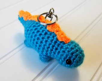 May include: A handmade crochet dinosaur keychain in bright blue with orange accents. The small dinosaur has a key ring attached, and a black bead for an eye. The keychain is on a white surface.