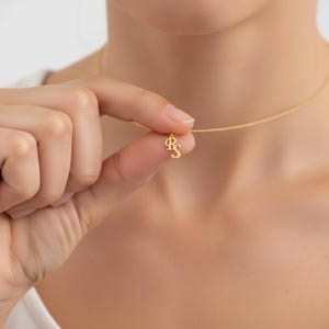 May include: A delicate gold-colored necklace with a pendant featuring the intertwined letters "R" and "S". The pendant hangs from a thin chain. The necklace is held up by a hand, showcasing the jewelry against a neutral background.