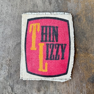 May include: A rectangular fabric patch with a frayed edge featuring a red background enclosed by a black border. The text Thin Lizzy is printed in a stylized yellow and black font, with the letters T and L appearing in yellow and the rest in black.