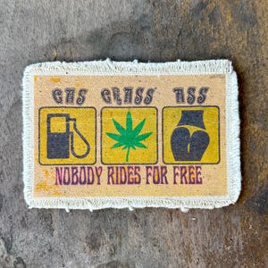 May include: A rectangular fabric patch with frayed edges featuring three yellow squares containing a black gas pump icon, a green cannabis leaf, and a black silhouette of buttocks. The text reads GAS GRASS ASS and NOBODY RIDES FOR FREE.