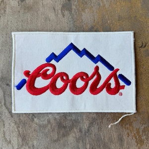 May include: A rectangular white fabric patch featuring an embroidered design. The design includes a blue mountain range graphic positioned above the red cursive text Coors. A small registered trademark symbol is visible next to the red text.