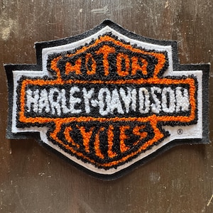 May include: A shield-shaped embroidered patch featuring the Harley-Davidson Motor Cycles logo. The design includes orange and white thread on a black background, with the text MOTOR, HARLEY-DAVIDSON, and CYCLES clearly visible in a bold, stylized font.