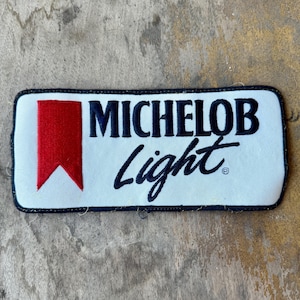May include: A rectangular white fabric patch with a black border. On the left is a red vertical banner shape with a pointed bottom. To the right, the text MICHELOB is embroidered in black block letters above the word Light in black script.