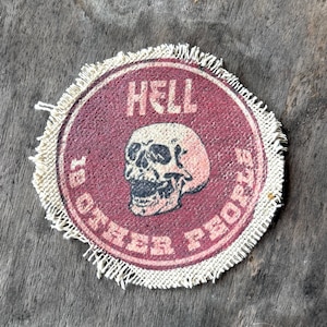 May include: A circular fabric patch with a frayed edge featuring a maroon background and a central illustration of a human skull. The text HELL IS OTHER PEOPLE is printed in a light cream color, with HELL at the top and IS OTHER PEOPLE below.