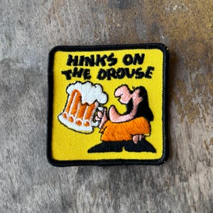 May include: A square embroidered patch with a yellow background and a black border. It features a cartoon character holding a large beer mug with white foam. The text "HINKS ON THE DROUSE" is embroidered in black letters at the top of the patch.