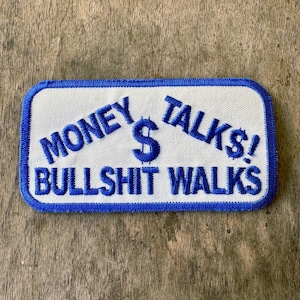 May include: A rectangular white patch with blue embroidered text that reads "MONEY TALKS! BULLSHIT WALKS" with a blue dollar sign. The patch has a blue border and is on a wooden surface.