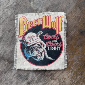 May include: A square fabric patch with frayed edges featuring a graphic of a wolf wearing a Coors Light cap. The text reads BeerWolf, Coors and Coors Light in yellow, red, and white lettering against a black circular background on a beige base.