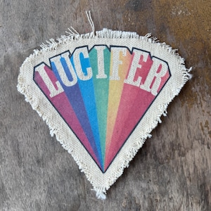 May include: A triangular fabric patch with frayed edges featuring a rainbow-colored sunburst design. The word LUCIFER is printed in large, bold white capital letters across the top of the colorful rays against a natural off-white background.