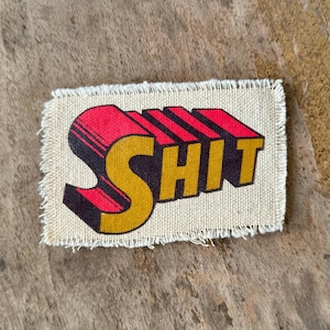May include: A rectangular fabric patch with frayed edges featuring the word SHIT printed in a bold, three-dimensional style. The letters are colored in a vibrant mustard yellow with red and black accents, set against a light beige canvas background.