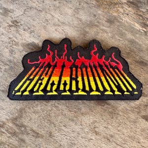 May include: Black embroidered patch featuring a stylized flame design in shades of red, orange, and yellow. The patch has a black border and is likely designed to be ironed or sewn onto clothing or accessories.