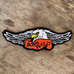May include: Embroidered patch featuring a white eagle with black and white wings, a yellow beak, and red eyes. The eagle is perched above a red banner with the words "BORN WILD" in black lettering. The patch has a black border.