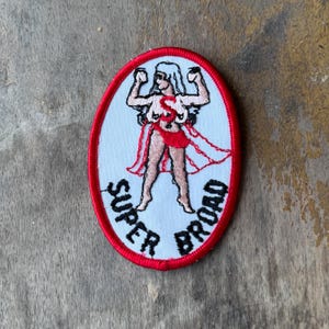 May include: An oval-shaped embroidered patch with a red border and a light blue background. It features a central figure with arms raised, wearing a red letter S on the chest and a red skirt. The text SUPER BROAD is stitched in black at the bottom.